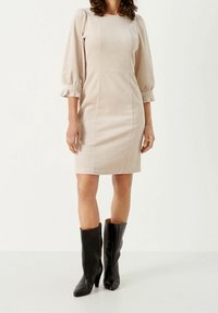 Robe en velours côtelé nude avec des manches bouffantes et des poignets volants, design ajusté, ourlet au genou, associée à des bottes noires à talons hauts montantes.