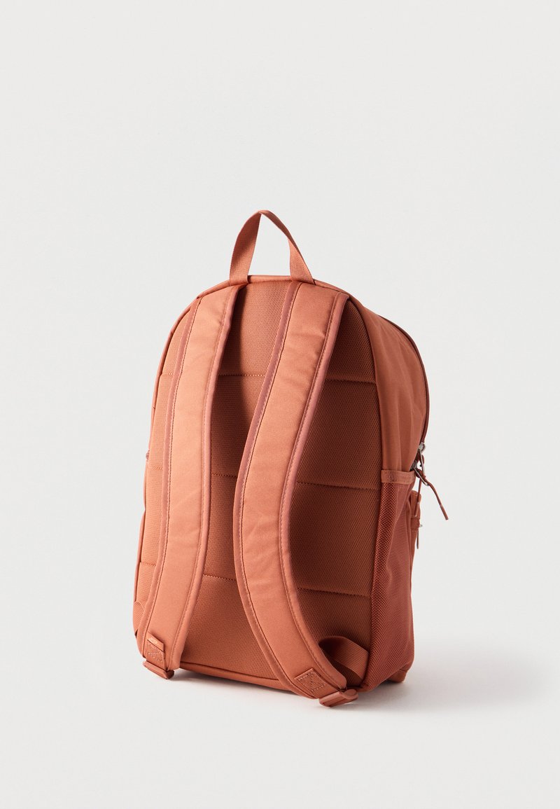 Mochila de color óxido con correas de hombro acolchadas, una asa superior y un bolsillo lateral con cremallera, mostrada sobre un fondo blanco liso.