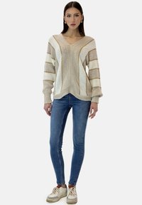 Suéter de punto con cuello en V en color beige con rayas color crema, que presenta un patrón texturizado. Combinado con jeans ajustados azules y zapatillas beige.