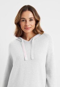 Lichtgrijze hoodie van zacht materiaal, met een voorzak, verstelbare koordjes en een label met witte en roze branding.