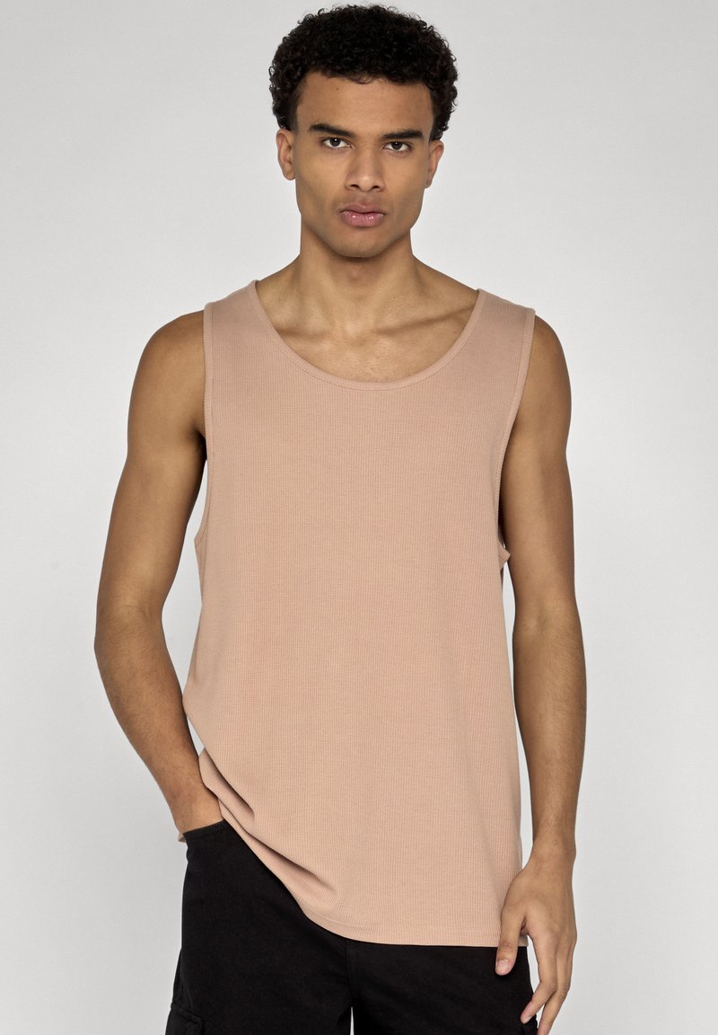 Jeune homme aux cheveux bouclés, portant un débardeur beige sans manches et un pantalon noir, debout devant un fond uni clair.