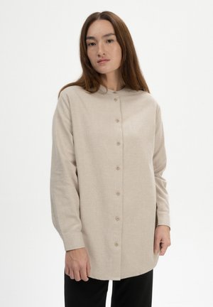 Frau mit langen braunen Haaren, die ein beiges, knöpfbares Hemd mit langen Ärmeln und eine schwarze Hose trägt und vor einem schlichten weißen Hintergrund steht.