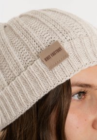 Crème gebreide beanie met ribbeltextuur, voorzien van een leren label met reliëftekst. Zacht, rekbaar materiaal met een gestructureerde bovenkant.