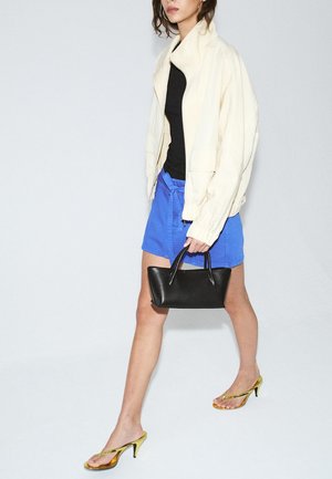 Femme marchant en veste blanche, haut noir, short bleu, portant un sac à main noir et des sandales dorées à talons hauts.
