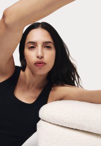 Zwarte tanktop op een individu met lang, donker haar, leunend op een gestructureerde witte kussen, tegen een effen witte achtergrond.