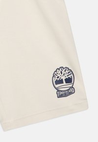 Camiseta de algodón beige con un logo redondo que presenta un diseño de un árbol y el texto "TIMBERLAND Est. 1973" en azul marino en la esquina inferior derecha.