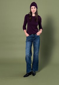 Femme debout les mains dans les poches, portant une chemise à manches longues violet foncé, un bonnet assorti, un jean bleu, et des bottines noires sur fond vert.