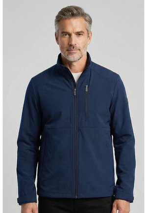 Homme aux cheveux gris et à la barbe, portant une veste bleu marine à fermeture éclair avec un col montant et une poche poitrine zippée, debout devant un fond uni.