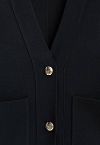 Black knitted cardigan with a V-neck, featuring two front pockets and gold-tone button accents. Smooth texture with ribbed detailing.