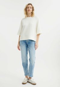 Biała, oversize bluza z krótkimi rękawami, zestawiona z jasnoniebieskimi dżinsami i beżowymi trampkami. Materiał wydaje się miękki i teksturowany.