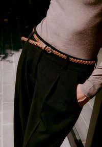 Braided tan leather belt worn over black high-waisted trousers, paired with a light gray long-sleeve top. Smooth texture and tailored fit.