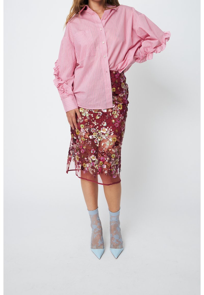 Femme portant un chemisier rose à rayures avec des manches volantées, une jupe transparente bordeaux brodée de fleurs, des chaussettes légères bleu clair à motifs, et des talons pointus.