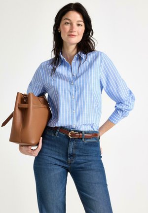 Kvinde med mørkt hår iført blå og hvide stribede skjorte med knapper og jeans, der holder en stor brun lædertaske på armen.