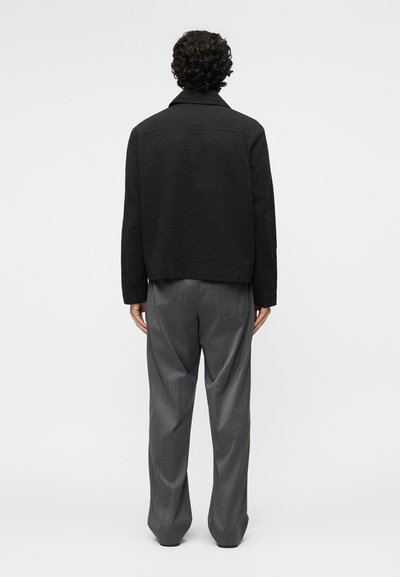 Göndör hajú személy háttal áll, texturált fekete dzsekit és laza, szürke csíkos nadrágot visel, egyszerű háttér előtt.