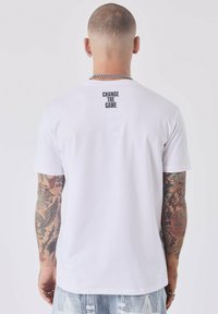 T-shirt bianco in cotone con scollatura a girocollo e maniche corte, con la scritta "CAMBIA IL GIOCO" stampata in nero sulla parte superiore della schiena.