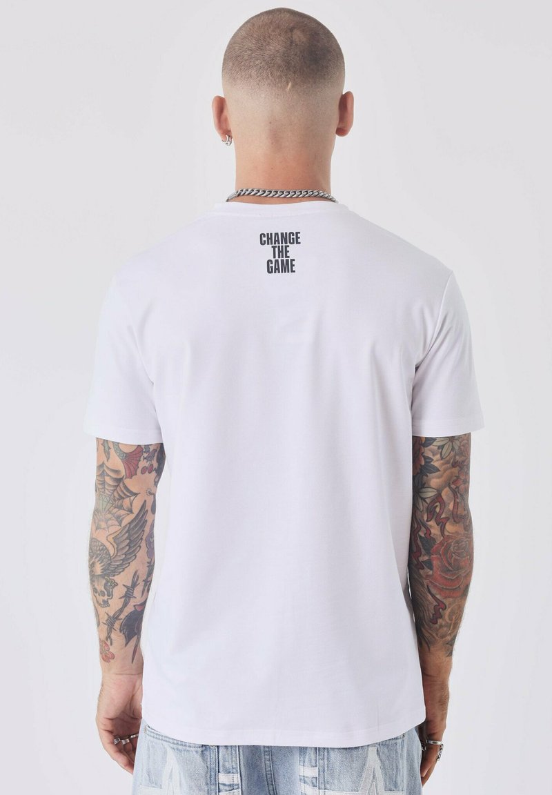 T-shirt bianco in cotone con scollatura a girocollo e maniche corte, con la scritta "CAMBIA IL GIOCO" stampata in nero sulla parte superiore della schiena.