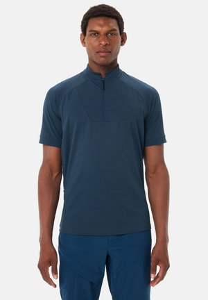 Mann trägt ein marineblaues, kurzärmeliges, zip-up Sportshirt und passende Hose, steht frontal vor einem weißen Hintergrund.