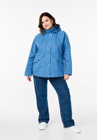 Giacca impermeabile azzurro chiaro con cappuccio, cordoncini regolabili, bottoni a pressione e due tasche frontali; abbinata a pantaloni in denim blu e scarpe bianche.