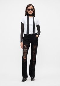Jeans negros desgastados con grandes desgarros, combinados con una camiseta blanca y una corbata de satén negra. El modelo lleva gafas de sol y elegantes botas negras.