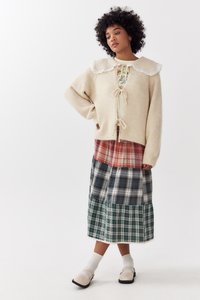 Vrouw met krullend haar draagt een beige gebreide cardigan met kantkraag, een gelaagde graphic tee, een patchwork geruite rok, witte sokken en beige Mary Jane schoenen.