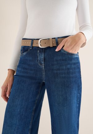 Jean en denim bleu à jambes larges, ceinturé d'une ceinture marron clair avec une boucle argentée et des clous décoratifs. Le haut est un t-shirt blanc ajusté à manches longues.