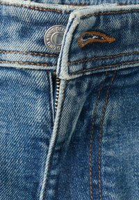 Zbliżenie na niebieskie dżinsy z rozpiętym zamkiem błyskawicznym, metalowym guzikiem oraz widocznym pomarańczowym przeszyciem na pasku i w okolicy zamka.