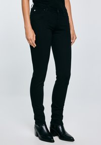 Fekete, szűk fazonú farmernadrágot és fekete, sarokkal ellátott bokacsizmát viselő személy, egyszínű világosszürke háttér előtt áll.