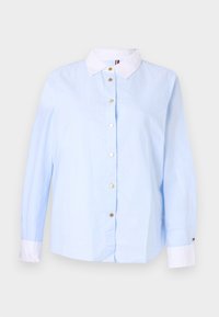 Blusa a maniche lunghe azzurro chiaro con colletto e polsini bianchi, bottoni dorati sul davanti e logo sottile sul polsino sinistro.