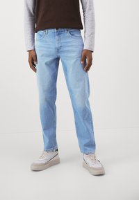 Jeans in denim azzurro chiaro con una vestibilità dritta, cinque tasche e leggere sfumature. Indossati con sneakers beige e bianche, in piedi su uno sfondo neutro.