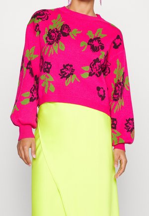 Femme portant un pull floral rose vif avec des fleurs vertes et noires assorti d'une jupe jaune néon, montrant le torse et les mains.