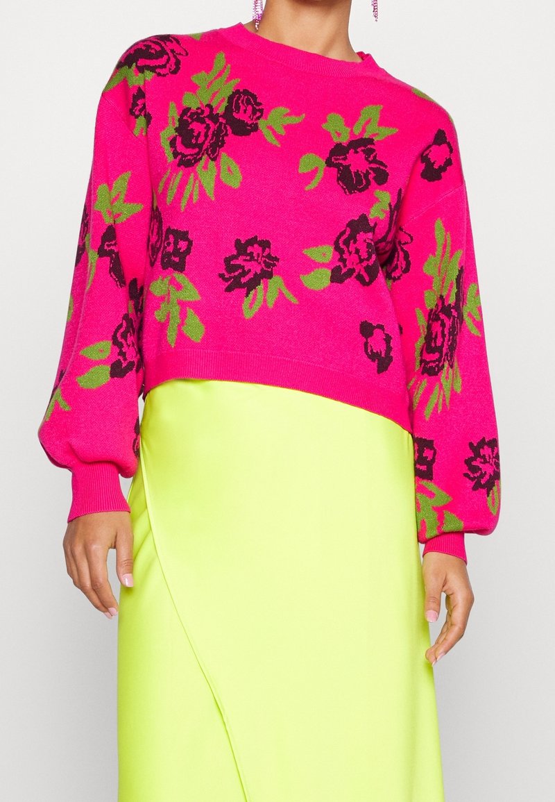 Femme portant un pull floral rose vif avec des fleurs vertes et noires assorti d'une jupe jaune néon, montrant le torse et les mains.