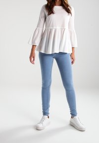 Weißes Oberteil mit Glockenärmeln und weit auslaufendem Saum, kombiniert mit hellblauen Skinny-Jeans und weißen Sneakers. Glatte Stoffqualität, lockere Passform.
