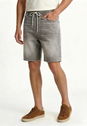 Muž nosi svetlosivé denimové šortky s bielou šnúrkou, bielu košeľu a hnedé semišové tenisky, stojí s jednou rukou v vrecku.