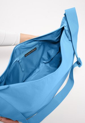 Sac à épaule bleu tenu ouvert montrant un intérieur spacieux avec une poche intérieure zippée et une étiquette en tissu portant la mention « From Trash To Treasure ».