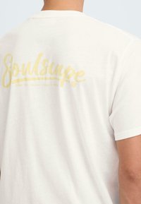T-shirt en coton blanc avec un logo "Soulscape" jaune en cursive au dos, accompagné du slogan "Brisez la routine - Sortez".