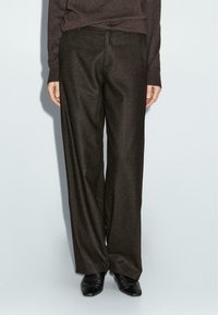 Pantalons larges marron foncé en tissu texturé, dotés d'une fermeture à l'avant et de poches, associés à des chaussures noires à talons pointus.