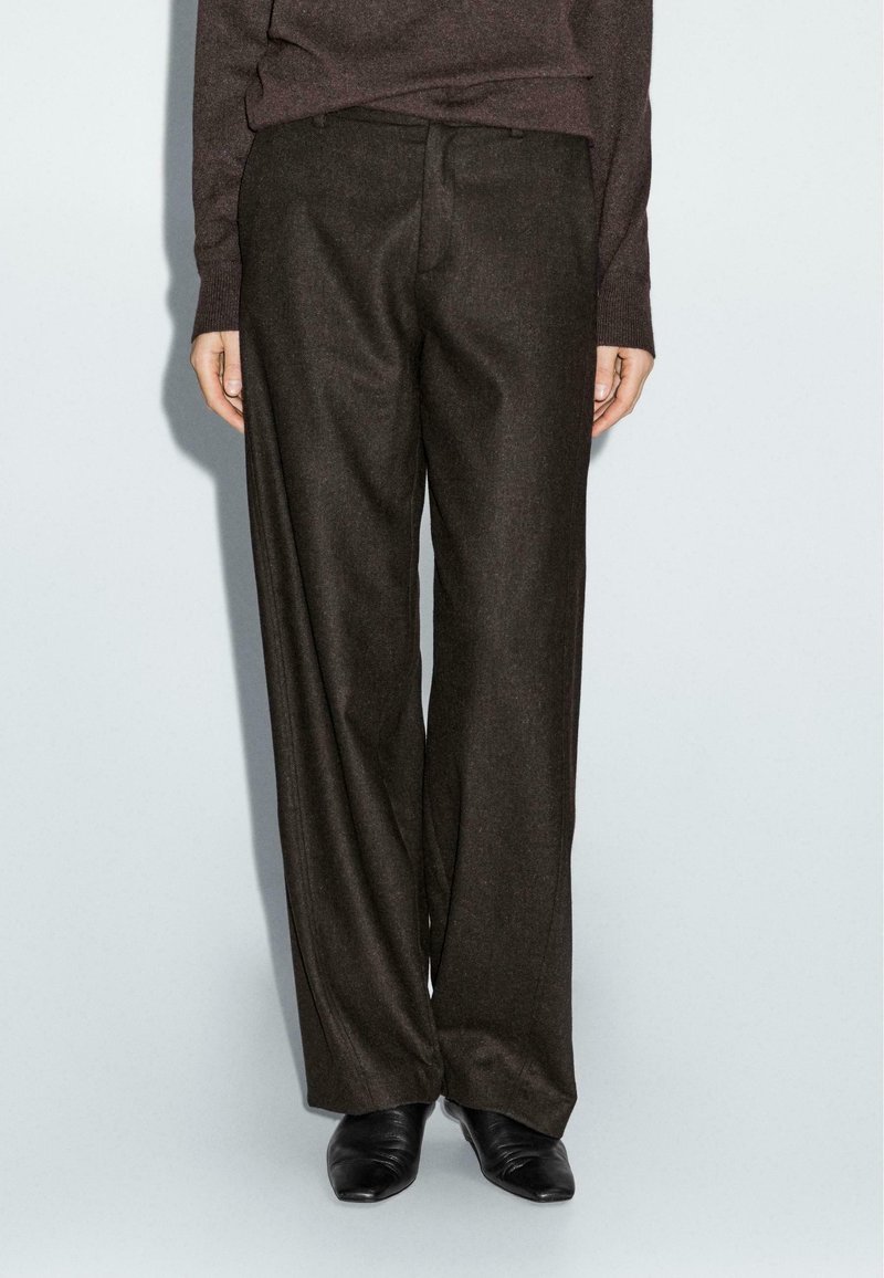 Pantalons larges marron foncé en tissu texturé, dotés d'une fermeture à l'avant et de poches, associés à des chaussures noires à talons pointus.