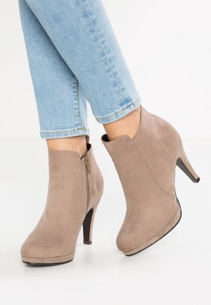 Botas de ante beige claro hasta el tobillo con cremalleras laterales y tacones altos, llevadas con jeans azul claro sobre un fondo blanco.