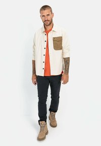 Lichtbeige button-up shirt met een bruine zak, oranje T-shirt, zwarte broek en tan boots. Tatoeages zichtbaar op de armen.