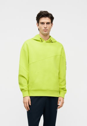 Ung mand iført limegrøn hættetrøje med frontlomme og marineblå bukser, stående mod en ensartet lys baggrund.