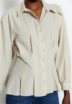 Jasnobeżowa koszula zapinana na guziki z długimi rękawami, szerokim kołnierzem i dopasowaną talią, noszona z niebieskimi dżinsami.