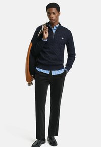 Pull en maille torsadée bleu marine avec zip, chemise rayée bleu clair en dessous, pantalon noir et chaussures noires. Mannequin tenant une veste marron.