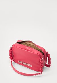 Rosa Kunstleder Crossbody-Tasche mit geflochtenen Akzenten, silbernem Logo, Reißverschluss und glatter Textur. Das Innere ist mit einem sandfarbenen Futter versehen.