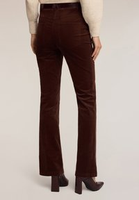 Pantalones de pana marrones con corte acampanado y bolsillos traseros, combinados con tacones burdeos brillantes. La tela tiene una textura suave y un ligero brillo.