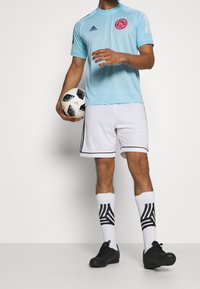 Hombre vestido con una camiseta de fútbol Ajax de color azul claro, pantalones cortos blancos, calcetines blancos con un patrón negro y botas negras, sosteniendo un balón de fútbol en blanco y negro.