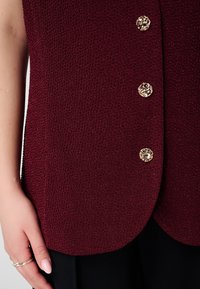 Bordeauxrode, gestructureerde mouwloze top met drie gouden decoratieve knopen en een afgeronde zoom, gecombineerd met zwarte stof aan de onderzijde.