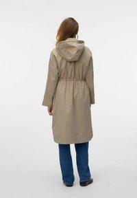 Beige regnrock med huva, elastisk midja, långa ärmar och knälång design. Buren över blå jeans och svarta skor.