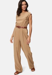 Combinaison beige avec des manches courtes, taille cintrée et une ceinture marron. Le tissu a une texture lisse ; le mannequin porte des sandales marron.