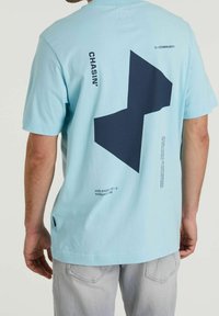 Jasnoniebieska bawełniana koszulka z dużym ciemnoniebieskim geometrycznym wzorem na plecach, z tekstem i grafiką w okolicy dekoltu i dołu.