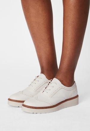 Personne à la peau foncée portant des chaussures brogues en cuir blanc texturé avec des semelles et des lacets beiges, se tenant sur une surface blanche.